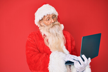Old senior man with grey hair and long beard wearing santa claus costume using laptop clueless and confused expression. doubt concept.