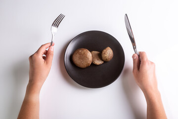 Raw Jersey cow mushrooms in a black plate and a knife and fork nearby on a white background top view. Human hands holding cutlery. Horizontal orientation. High quality photo