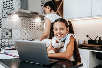 Young little girl having e-learning session during Covid-19 pandemic crisis lockdown or quarantine. Busy mother working in kitchen and preparing lunch in the background. 