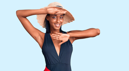 Young african american woman wearing swimsuit and summer hat smiling cheerful playing peek a boo with hands showing face. surprised and exited