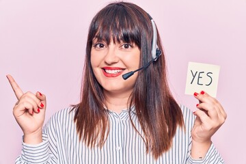 Young plus size woman wearing call center agent headset holding yes reminder smiling happy pointing...