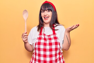 Young plus size woman wearing apron holding wooden spoon celebrating achievement with happy smile and winner expression with raised hand