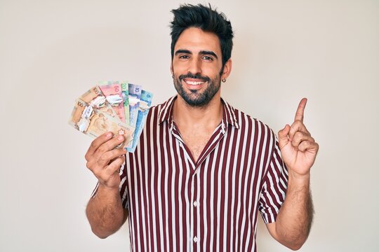 Handsome hispanic man with beard holding canadian dollars smiling happy pointing with hand and finger to the side