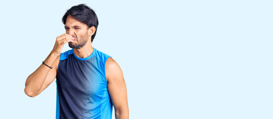 Handsome hispanic man wearing sportswear smelling something stinky and disgusting, intolerable...
