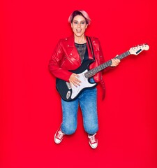 Young beautiful guitarist woman with pink short hair playing electric guitar smiling happy. Jumping with smile on face over isolated red background