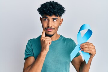 Young arab handsome man holding blue ribbon serious face thinking about question with hand on chin,...