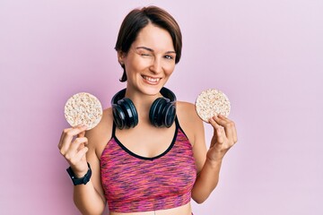 Young brunette woman with short hair wearing sportswear holding rice cakes winking looking at the...
