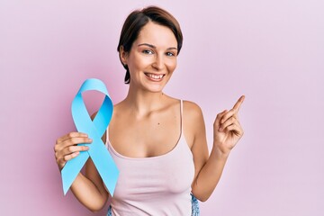 Young brunette woman with short hair holding blue ribbon smiling happy pointing with hand and finger to the side