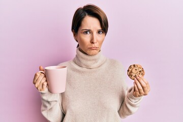 Young brunette woman with short hair drinking a cup of coffee and cookie depressed and worry for distress, crying angry and afraid. sad expression.