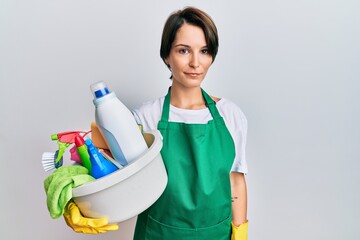 Young brunette woman with short hair wearing apron holding cleaning products relaxed with serious expression on face. simple and natural looking at the camera.