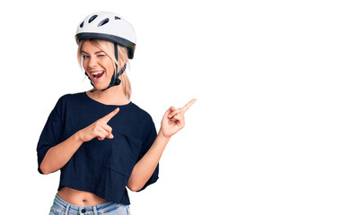 Young beautiful blonde woman wearing bike helmet smiling and looking at the camera pointing with two hands and fingers to the side.