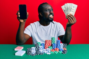Handsome young black man playing poker holding smartphone and dollars angry and mad screaming frustrated and furious, shouting with anger. rage and aggressive concept.