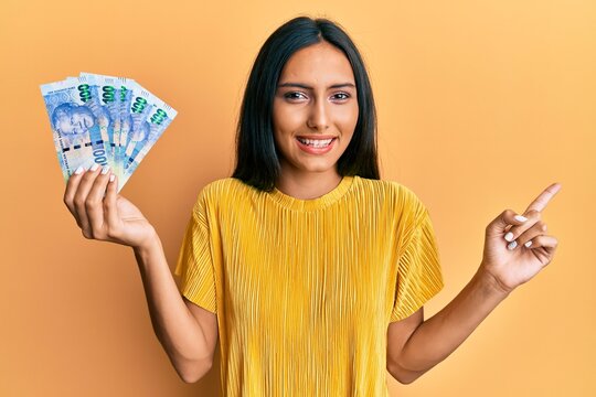 Young brunette woman holding south african 100 rand banknotes smiling happy pointing with hand and finger to the side