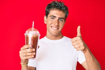 Young handsome man holding glass of smoothie smiling happy and positive, thumb up doing excellent and approval sign