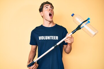 Young handsome man recycling plastic bottle holding litter pick up tool angry and mad screaming frustrated and furious, shouting with anger looking up.