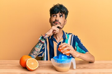 Young hispanic man sitting on the table using juicer serious face thinking about question with hand...