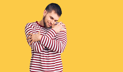 Young handsome man wearing striped sweater hugging oneself happy and positive, smiling confident. self love and self care