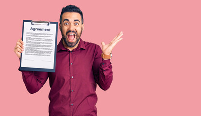 Young hispanic man holding clipboard with agreement document celebrating victory with happy smile and winner expression with raised hands