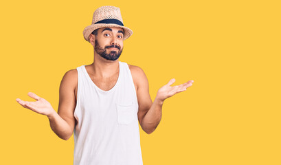 Young hispanic man wearing casual summer hat clueless and confused expression with arms and hands...