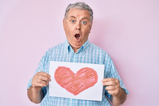 Senior grey-haired man holding heart draw afraid and shocked with surprise and amazed expression, fear and excited face.