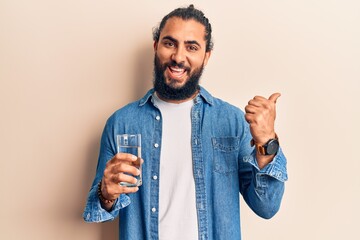 Young arab man drinking glass of water pointing thumb up to the side smiling happy with open mouth