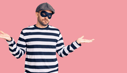 Young handsome man wearing burglar mask clueless and confused expression with arms and hands raised. doubt concept.