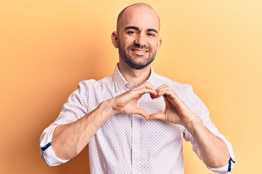 Young handsome bald man wearing elegant shirt smiling in love doing heart symbol shape with hands. romantic concept.