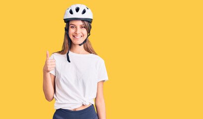 Young beautiful girl wearing bike helmet smiling happy and positive, thumb up doing excellent and approval sign