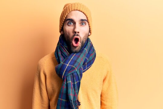 Young handsome man with beard wearing wool cap and scarf scared and amazed with open mouth for surprise, disbelief face