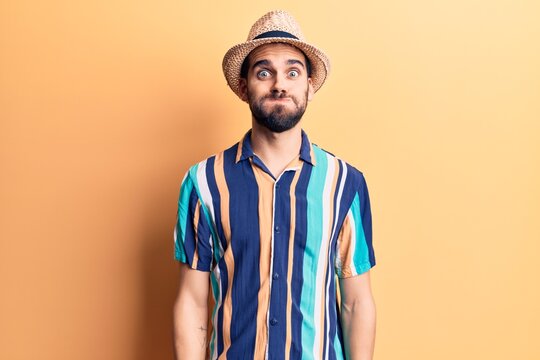 Young handsome man with beard wearing summer hat and shirt puffing cheeks with funny face. mouth inflated with air, crazy expression.