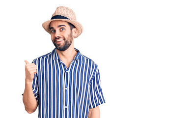 Young handsome man with beard wearing summer hat and striped shirt smiling with happy face looking and pointing to the side with thumb up.