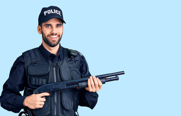 Young handsome man with beard wearing police uniform holding shotgun looking positive and happy...
