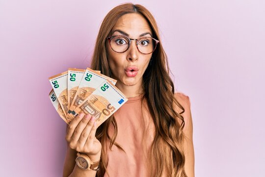 Young brunette woman holding bunch of 50 euro banknotes scared and amazed with open mouth for surprise, disbelief face