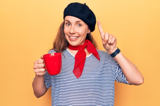 Young beautiful blonde woman wearing fashion beret drinking a cup of coffee smiling with an idea or question pointing finger with happy face, number one