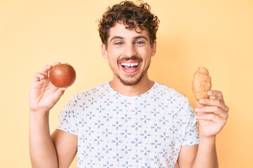 Young caucasian man with curly hair holding apple and croissant smiling and laughing hard out loud because funny crazy joke.