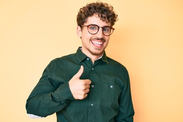 Young caucasian man with curly hair wearing casual clothes and glasses doing happy thumbs up...