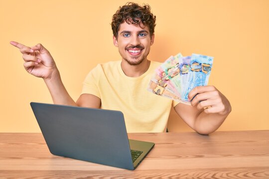 Young caucasian man with curly hair sitting on the table working with laptop and holding canadian dollars banknotes smiling happy pointing with hand and finger to the side