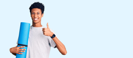 Young african american man holding yoga mat smiling happy and positive, thumb up doing excellent and approval sign