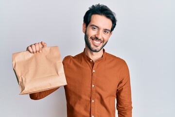 Young hispanic man holding take away food looking positive and happy standing and smiling with a confident smile showing teeth