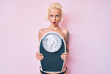 Young blonde woman with tattoo standing shirtless holding weighing machine in shock face, looking skeptical and sarcastic, surprised with open mouth