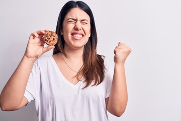 Young beautiful brunette woman eating chocolate cookie over isolated white background screaming proud, celebrating victory and success very excited with raised arm