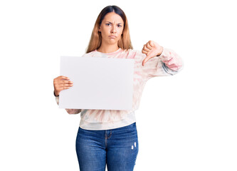 Beautiful young woman holding blank empty banner with angry face, negative sign showing dislike with thumbs down, rejection concept