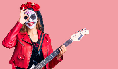 Woman wearing day of the dead costume playing electric guitar smiling happy doing ok sign with hand on eye looking through fingers