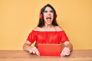 Brunette teenager girl using touchpad wearing headphones angry and mad screaming frustrated and furious, shouting with anger looking up.