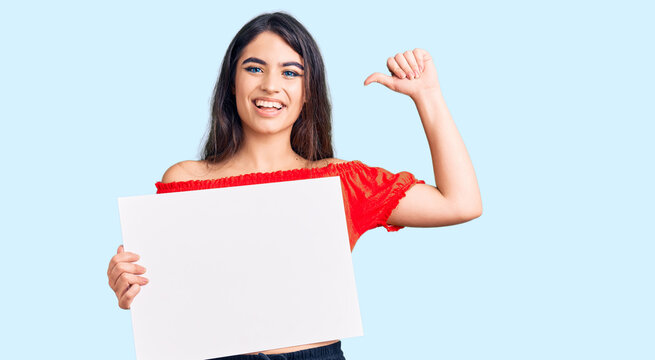 Brunette teenager girl holding blank empty banner pointing finger to one self smiling happy and proud