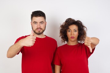 Young beautiful couple wearing red t-shirt on white background showing thumbs up and thumbs down, difficult choose concept - obrazy, fototapety, plakaty