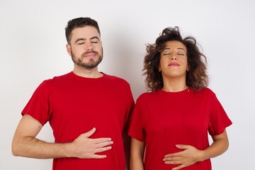 Fototapeta premium Young beautiful couple wearing red t-shirt on white background touches tummy, smiles gently, eating and satisfaction concept.