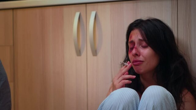 Traumatised wife smoking and crying sitting on the floor. Abused terrified beaten woman covered in bruises suffering injury from alcoholic violent brutal aggressive husband screaming.