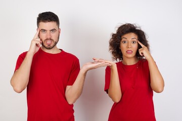 Young beautiful couple wearing red t-shirt on white background confused and annoyed with open palm showing copy space and pointing finger to forehead. Think about it.