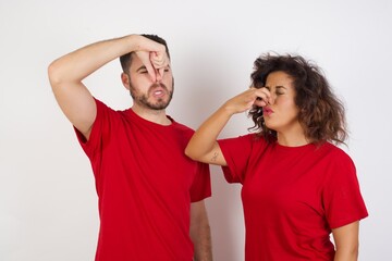 Young beautiful couple wearing red t-shirt on white background smelling something stinky and disgusting, intolerable smell, holding breath with fingers on nose. Bad smell